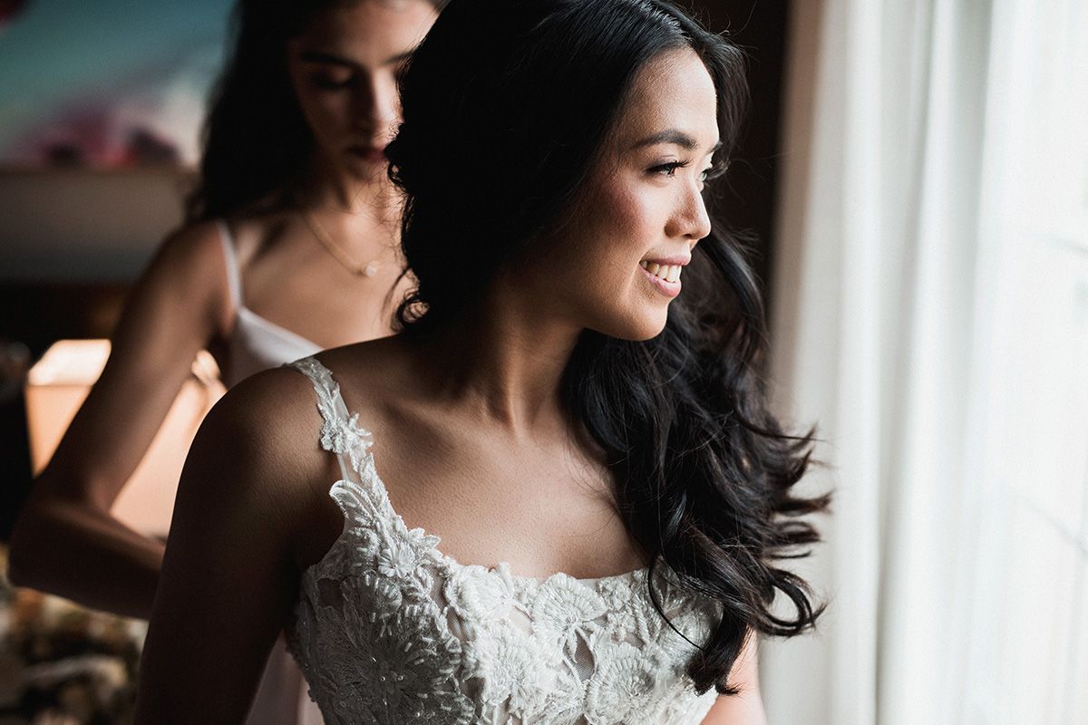 A bride in a lace wedding dress looks out a window with soft natural light illuminating her silhouette.
