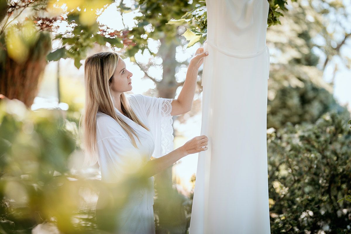 A bride-to-be inspects her wedding dress  hanging in a tree in a garden setting with soft sunlight filtering through trees.