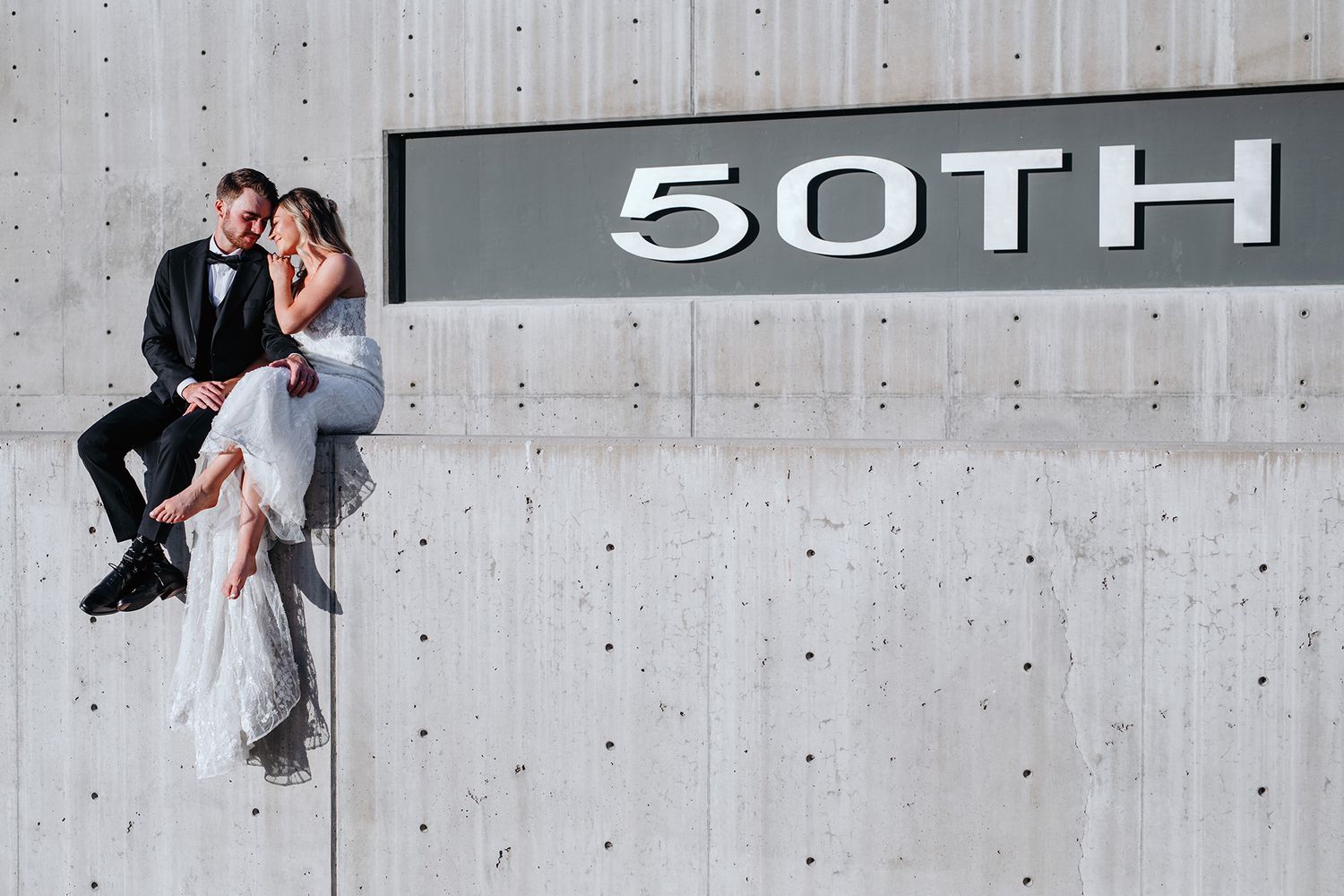 A couple in wedding attire poses against a modern concrete wall with the sign 50TH PARALLEL displayed in the background.