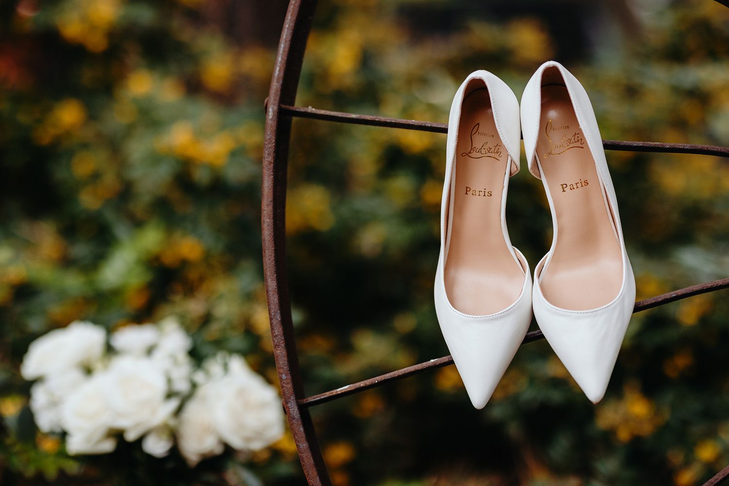 White wedding heels rest elegantly on a metal railing against a blurred garden background with yellow flowers.