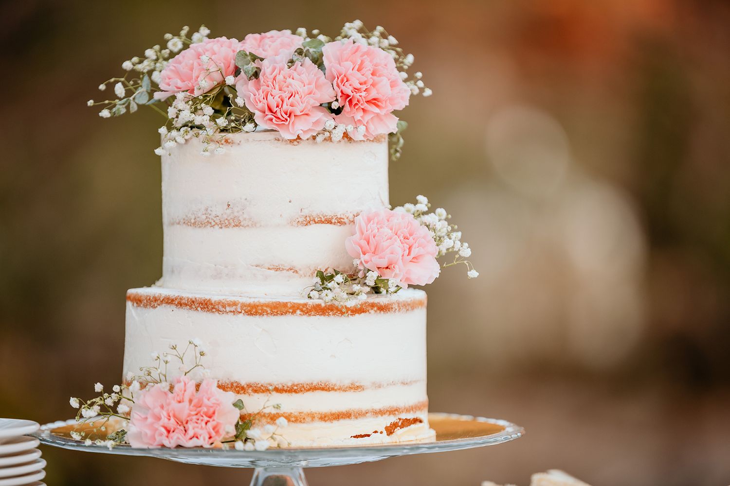A rustic naked wedding cake decorated with delicate pink carnations and baby's breath on a white cake stand.
