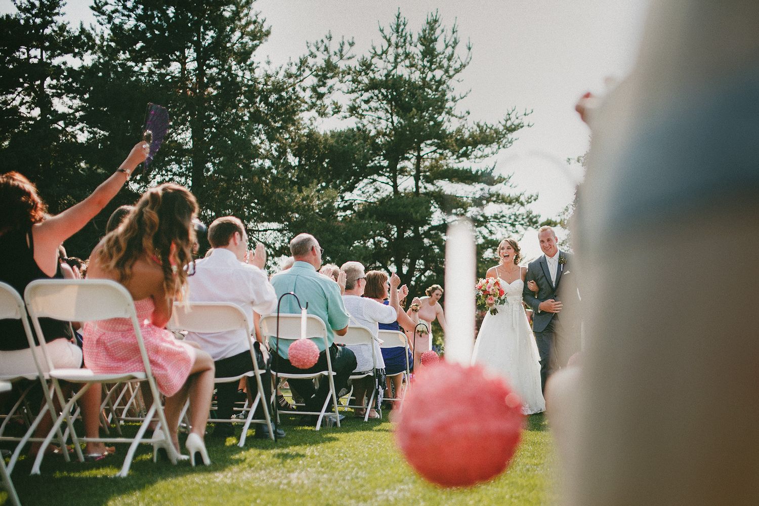 Wedding guests seated in white folding chairs on a lawn watch a ceremony while a red balloon floats in the foreground.