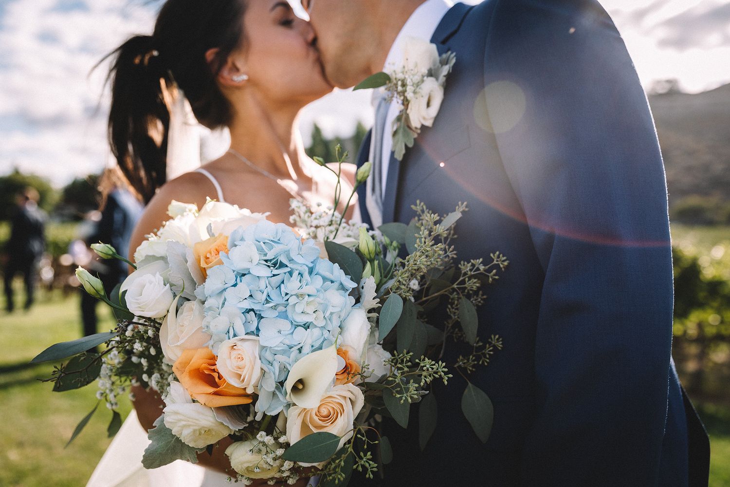 A romantic wedding couple embraces outdoors while holding a beautiful peach and white floral bouquet at sunset.