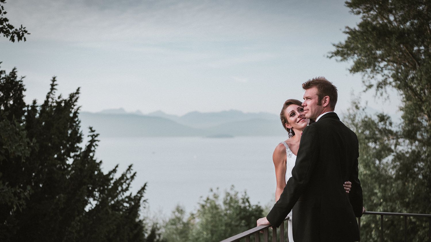 A couple in formal wedding attire embrace while overlooking a misty mountain lake surrounded by evergreen trees.