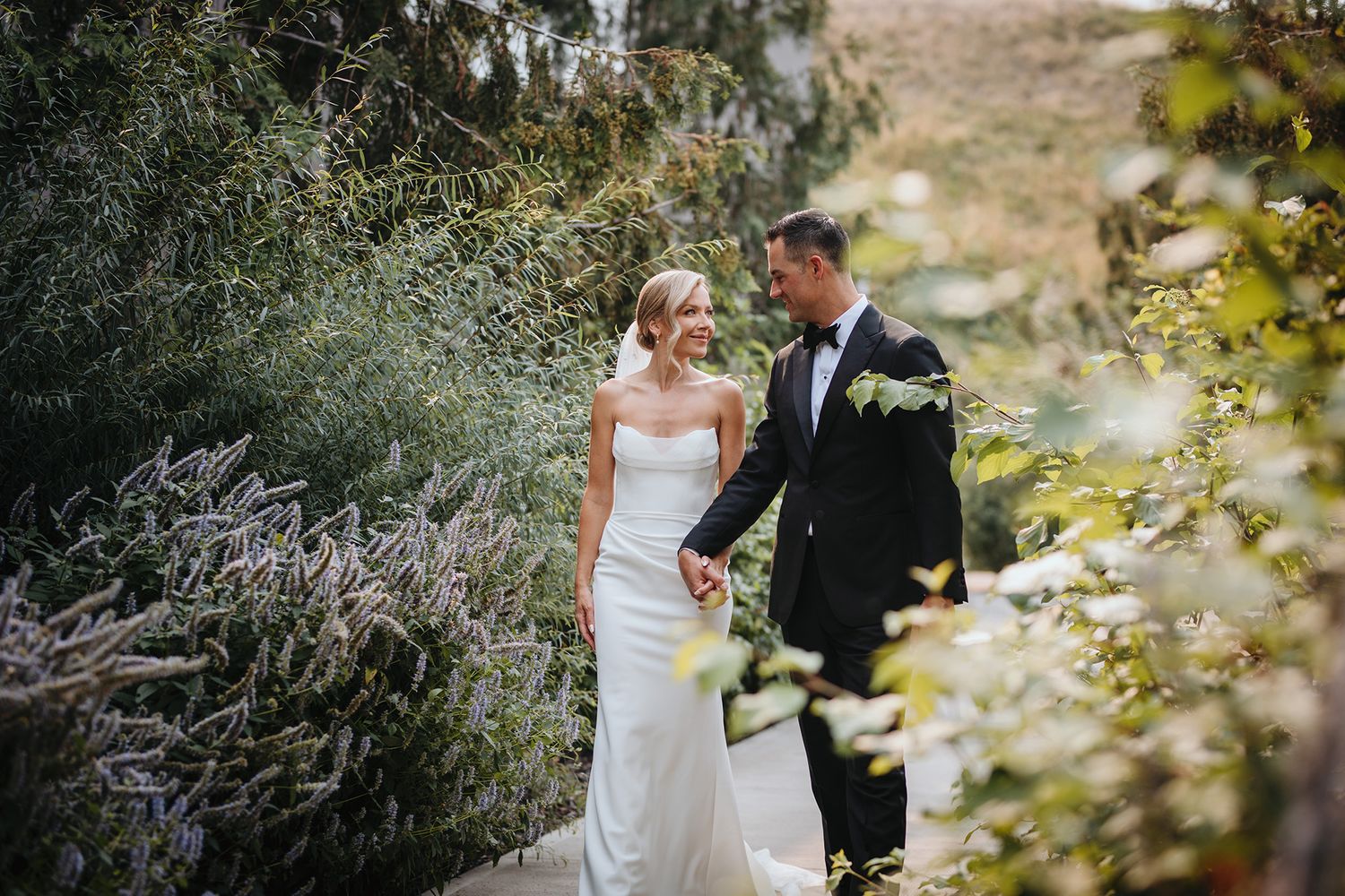 A couple in wedding attire embraces while walking in a lush outdoor garden setting at Cedar Creek Winery in Kelowna.