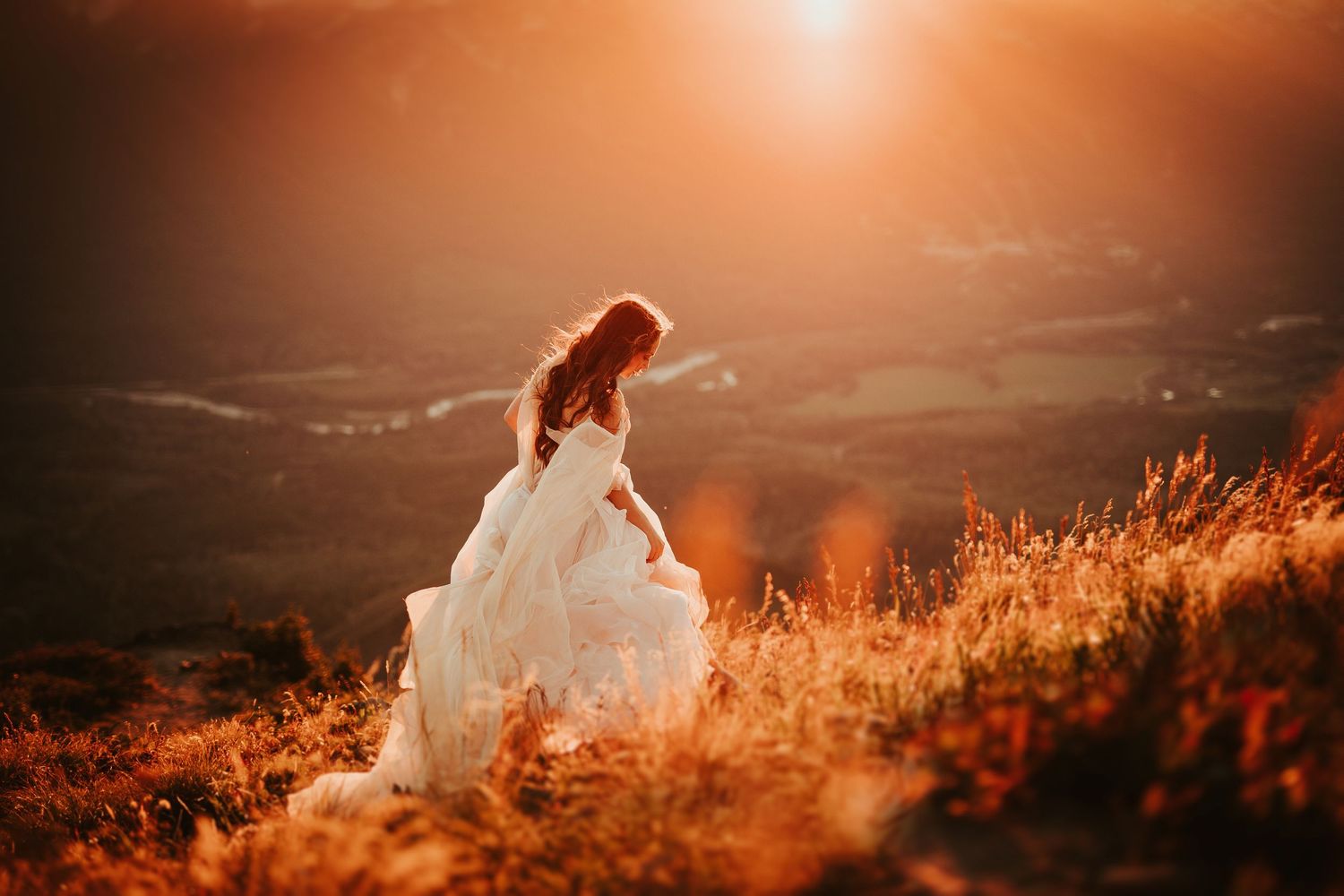Silhouetted bride in white dress stands on hilltop during golden sunset with glowing backlight.