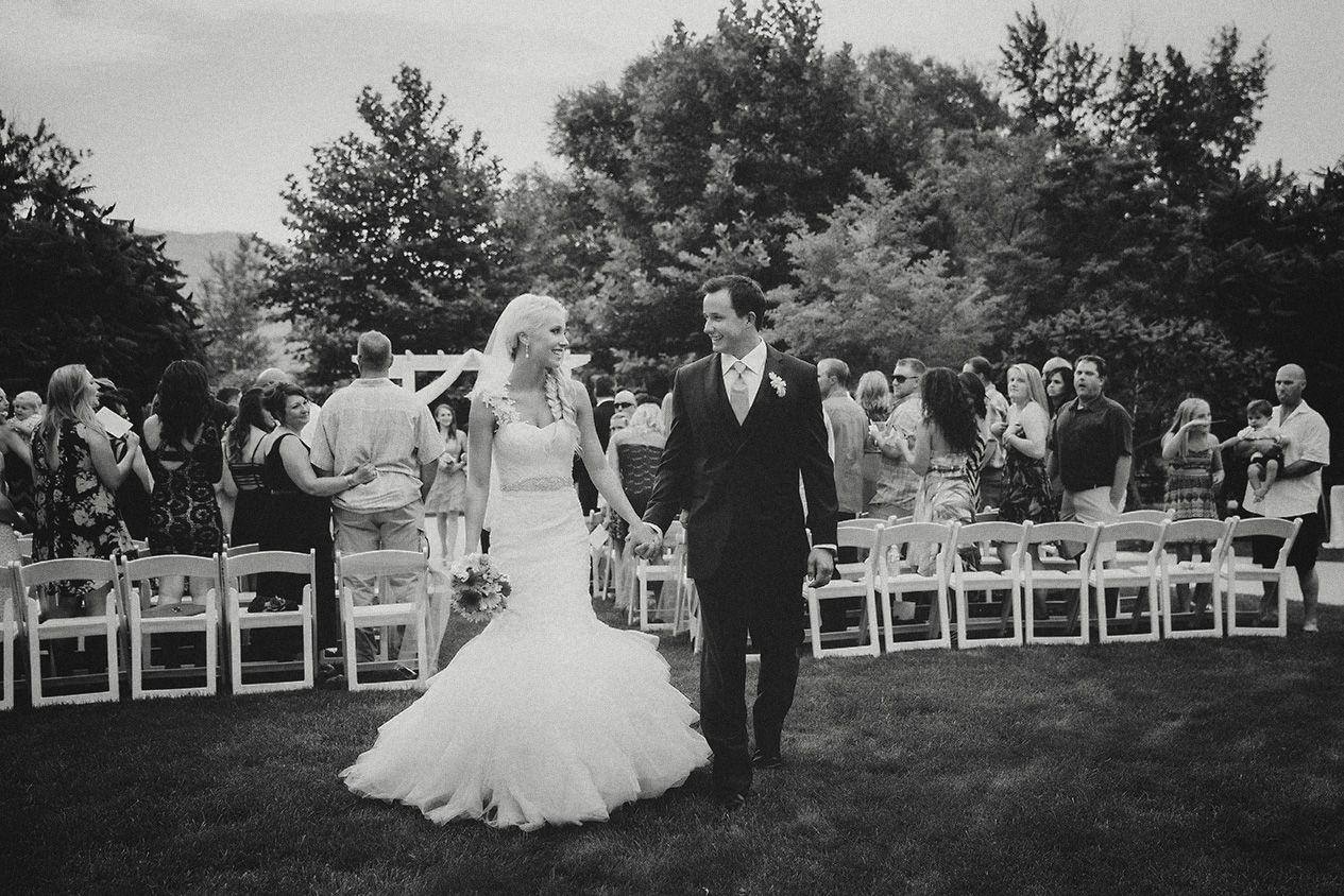 Black and white panoramic photo of an outdoor wedding ceremony with guests seated in white chairs on a lawn.