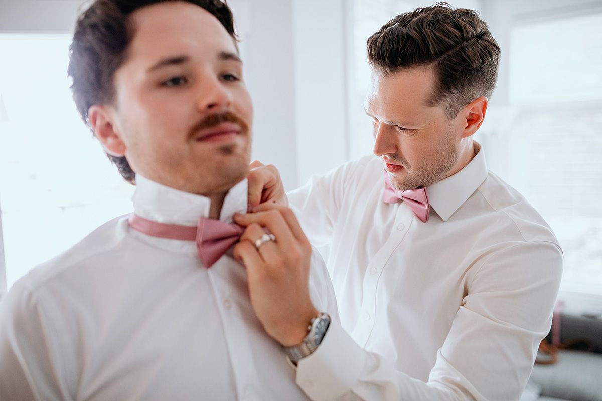 Two groomsmen help each other tie their pink bow-ties.