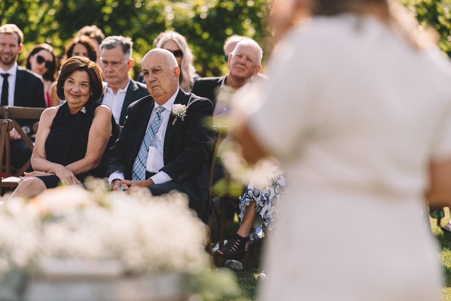 Wedding guests seated outdoors in white chairs during a garden ceremony on a sunny summer day.