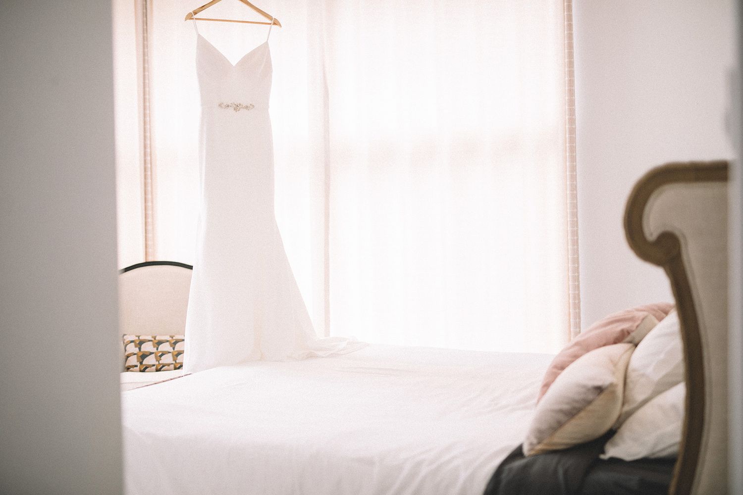 A white wedding dress hangs in a bright room with floor to cieling windows.