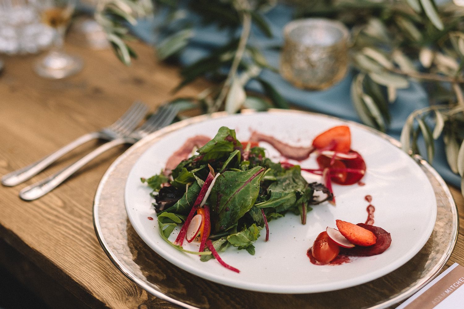 A fresh salad plate with mixed greens, prosciutto, and strawberries served on white dishes with olive branches nearby.