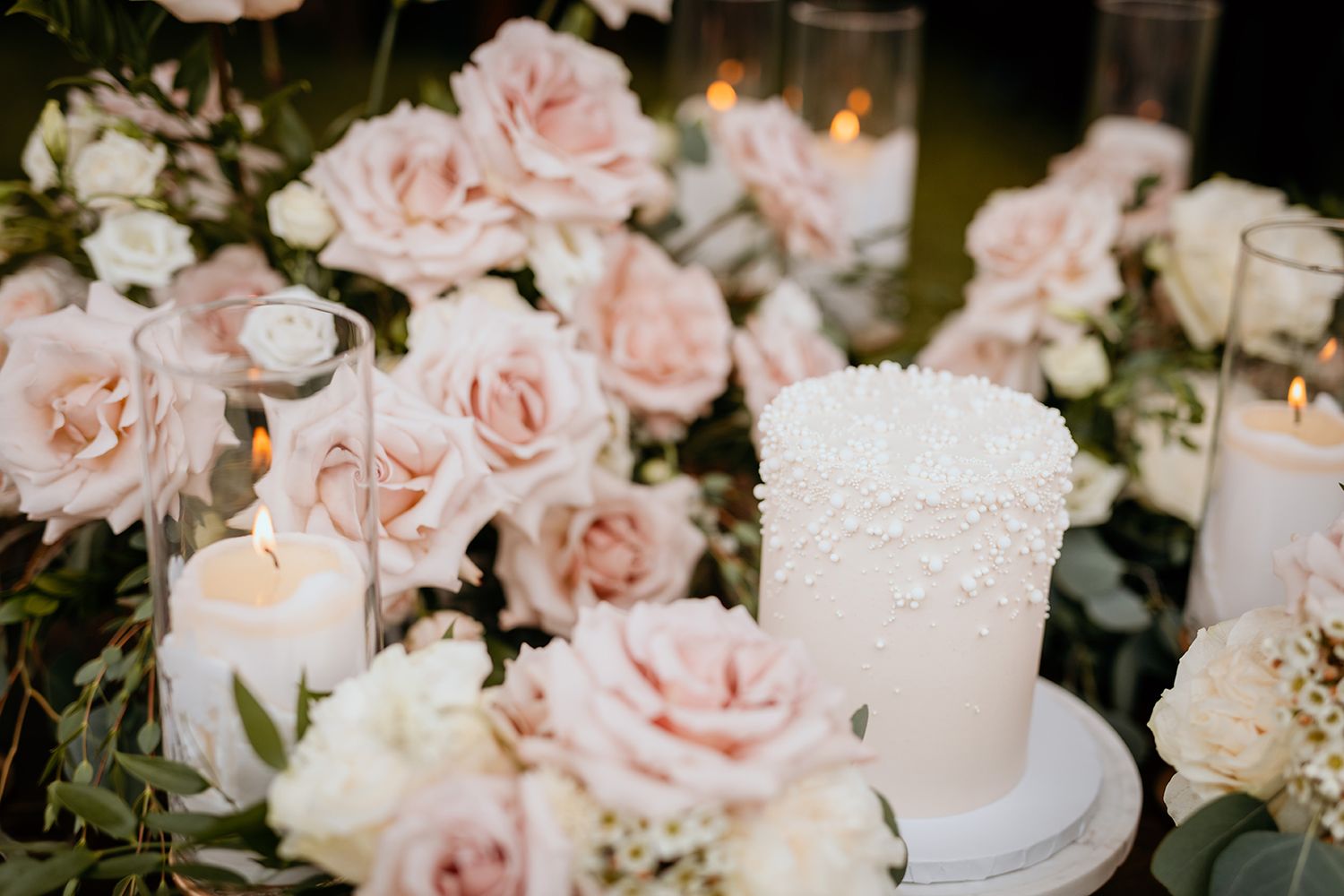 Elegant white wedding cake surrounded by soft pink roses and glowing candles in romantic evening setting.