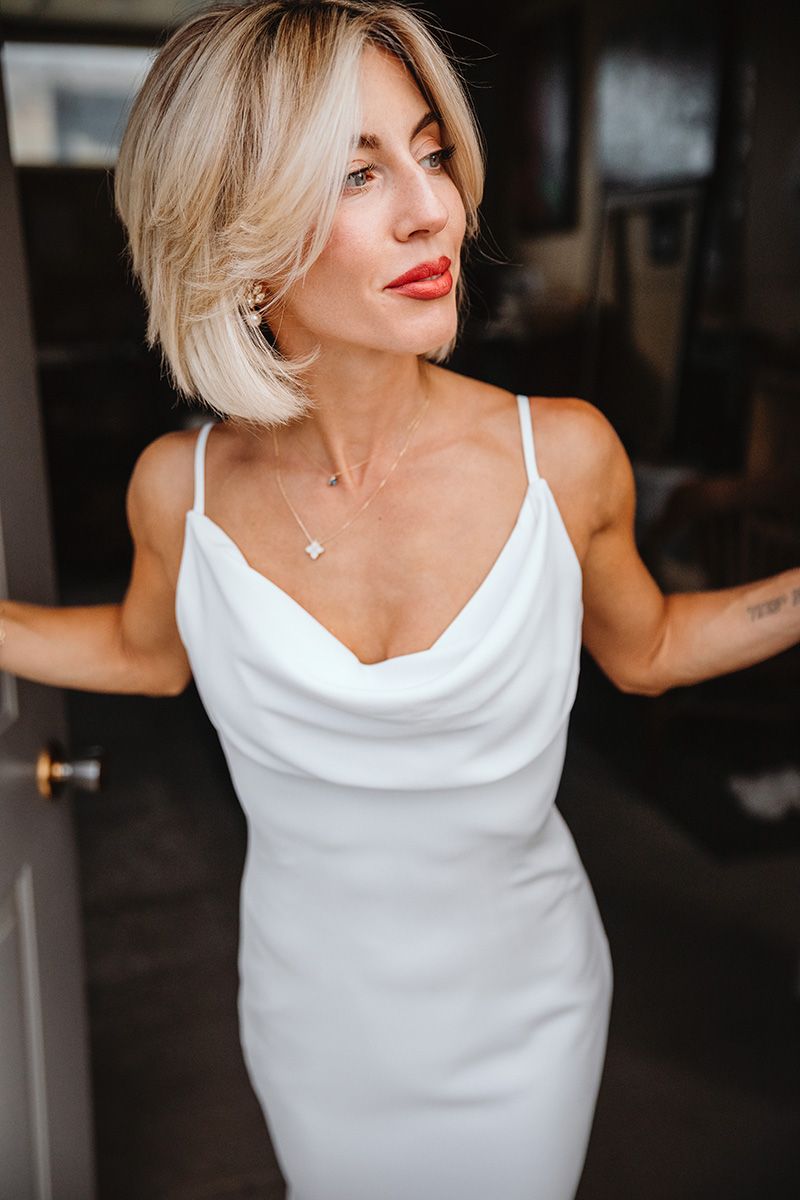A bride in a white sleeveless dress poses in the doorway of her bridal suite. She peers out toward her garden ceremony.