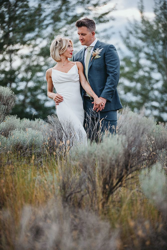 A romantic wedding couple stands together in sage brush with mountain scenery and evergreen trees in the background.