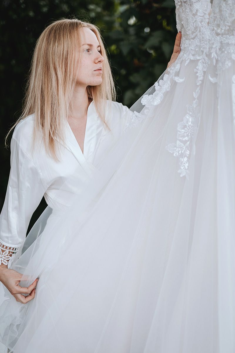 Woman in white robe examines delicate lace details on flowing white wedding dress.