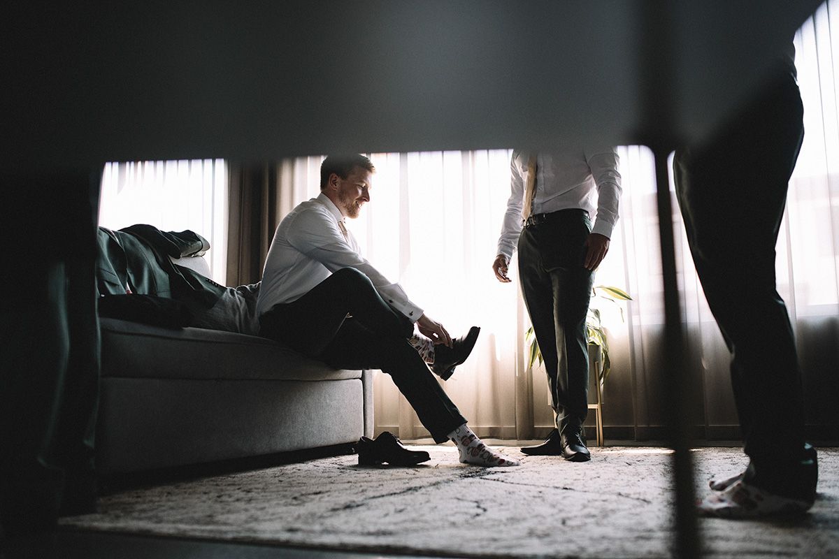 From a low angle through blurry foreground objects, a man seated on a couch, puts on his shoes while another man looks on.