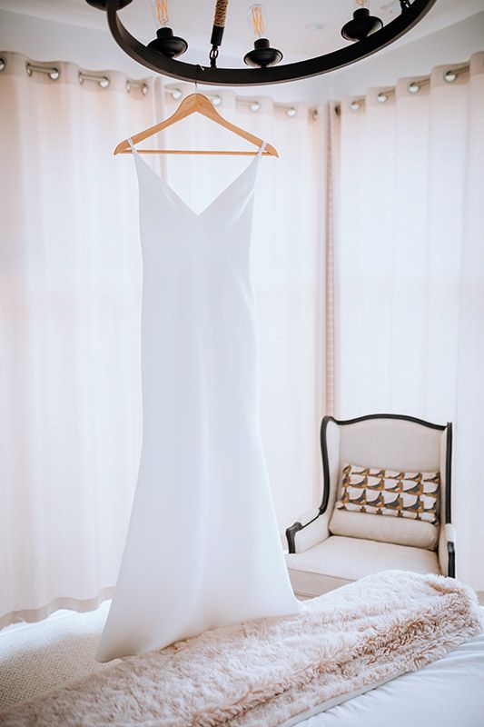 A white wedding dress hangs on a wooden hanger in a bright room with white curtains and elegant decor.