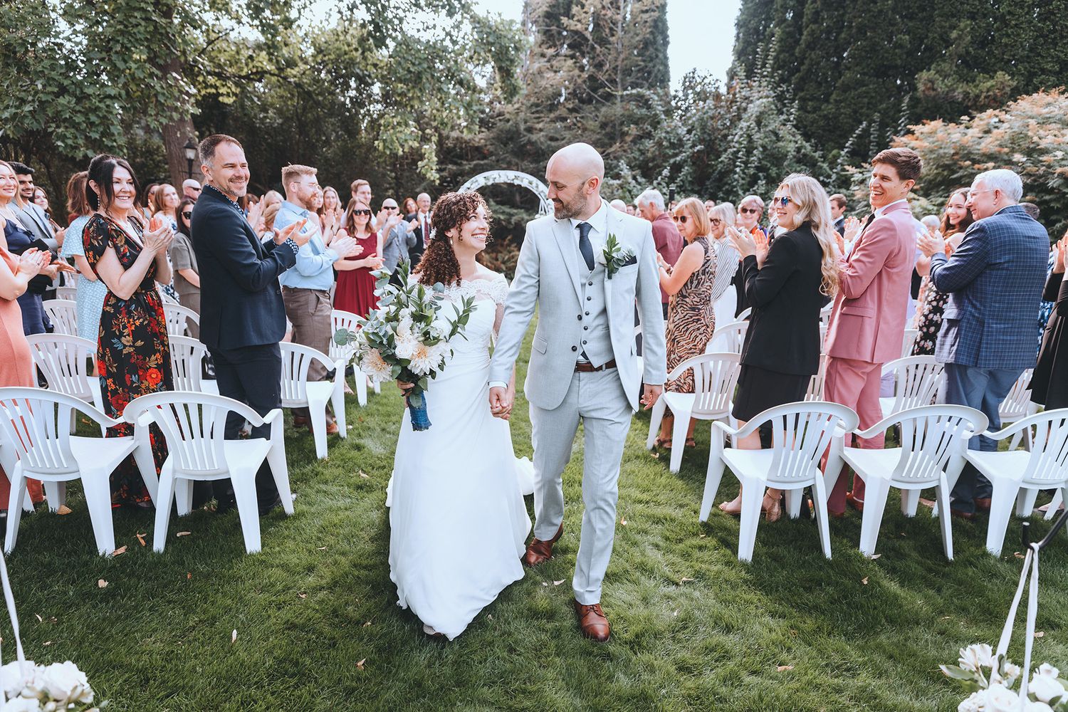 Newlywed couple walks down outdoor aisle between rows of white chairs as wedding guests celebrate and take photos.