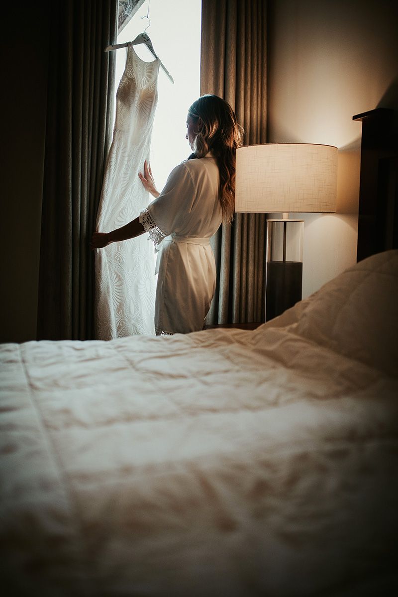 A dimly lit bride stands by window curtains in hotel room with soft morning light filtering through. She inspects her wedding dress.
