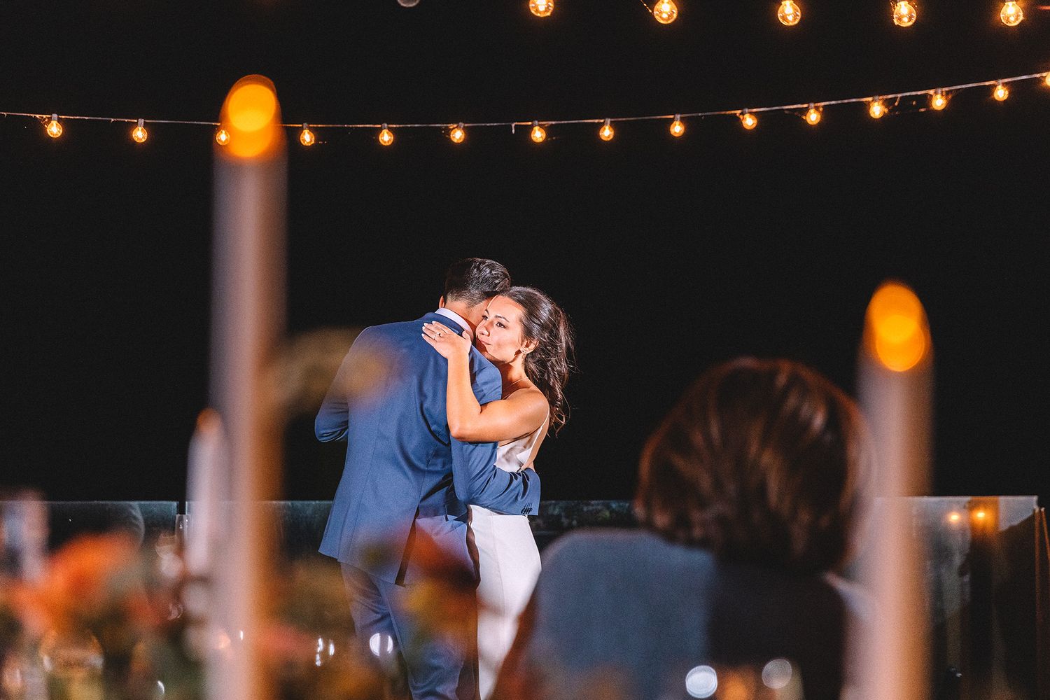 A couple shares a romantic dance under twinkling string lights at an outdoor evening celebration.