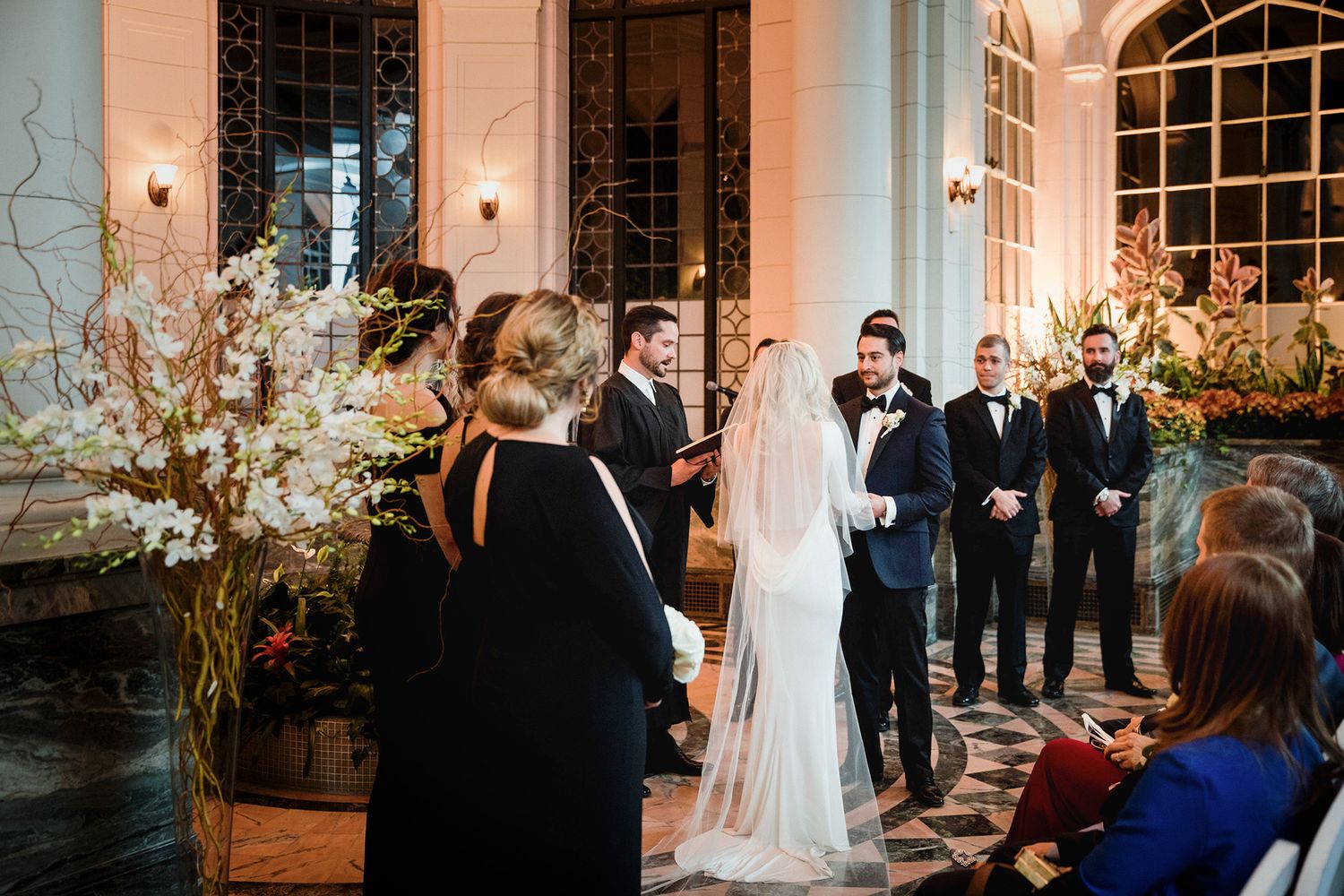 Wedding party stand in an elegant ballroom with arched windows and warm lighting during an evening celebration.