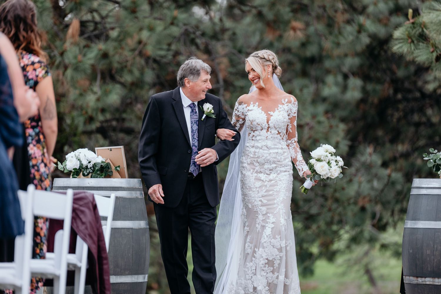 A bride and her father walk together on a garden path surrounded by lush greenery during her outdoor wedding ceremony.