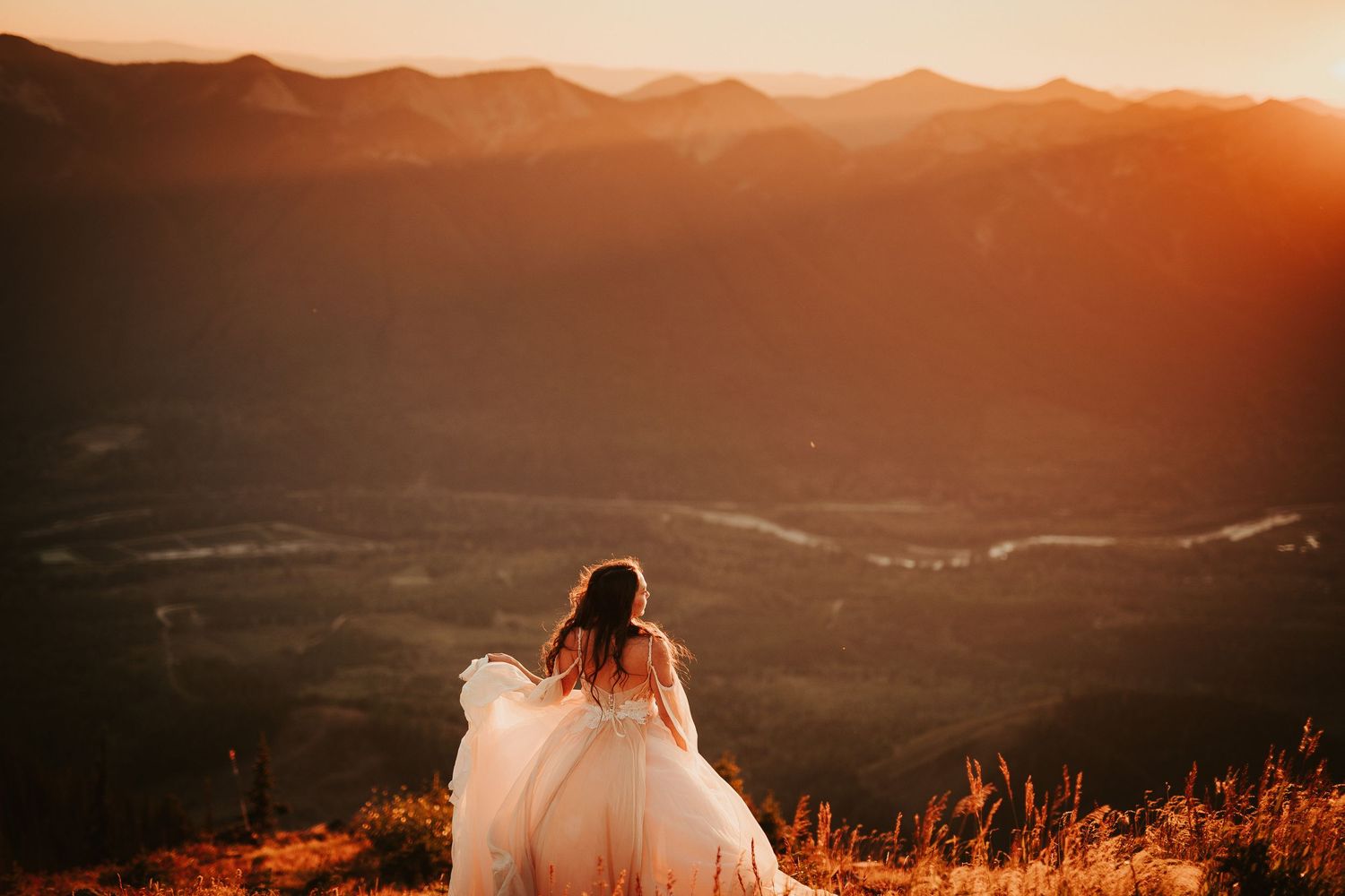 A bride in a flowing white dress stands on a hilltop overlooking a mountainous landscape at golden sunset.