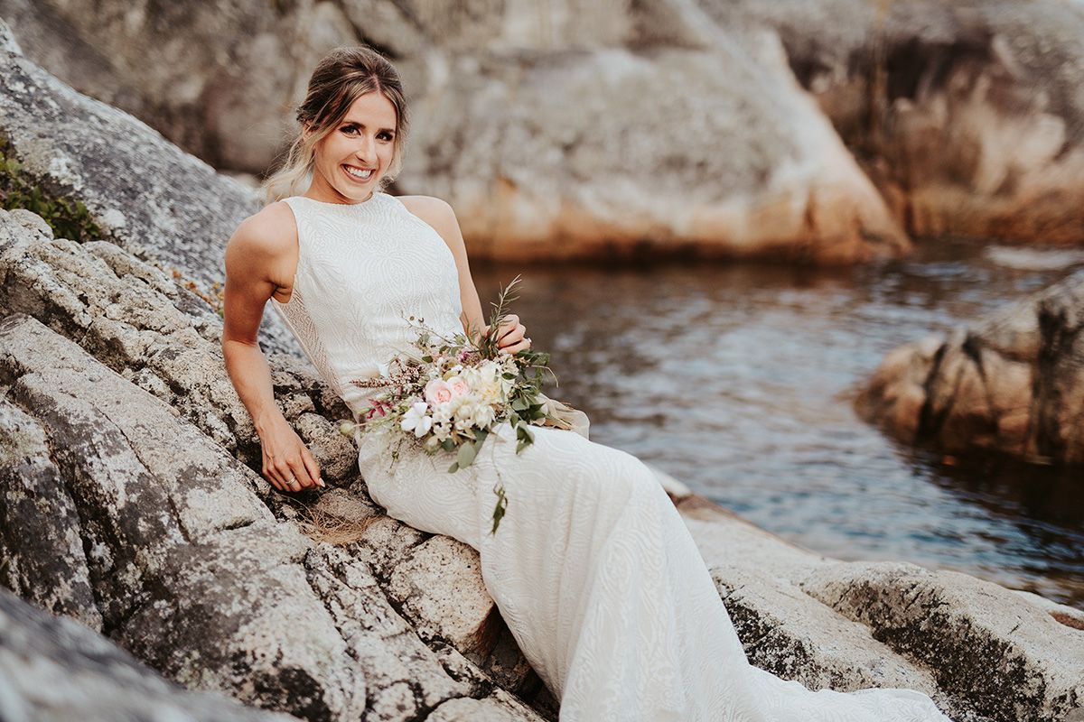 A bride in a white wedding dress poses gracefully on rocky coastal terrain near the ocean.