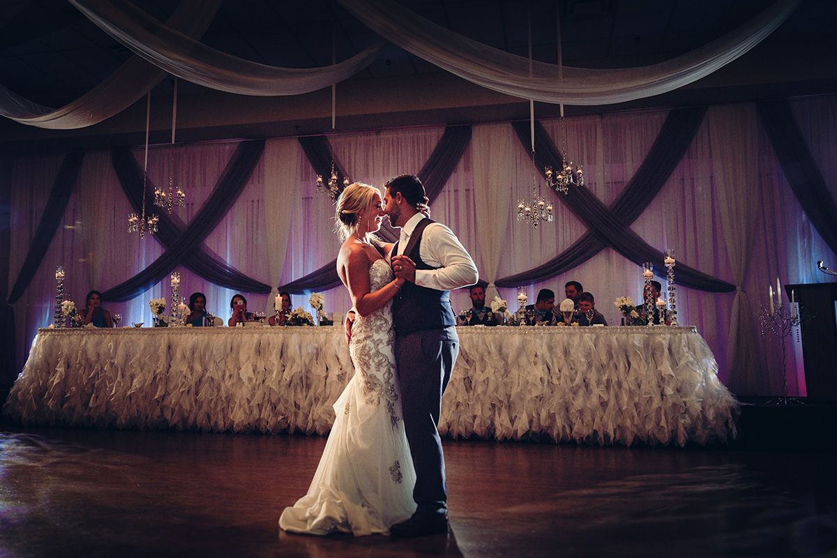 A romantic couple dances together during their wedding reception under elegant draped fabric decor and soft lighting.