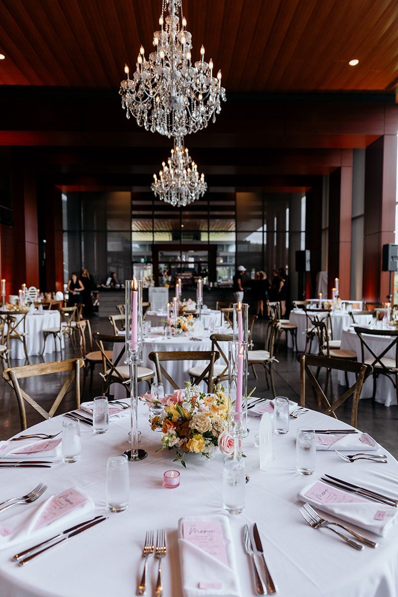 Elegant wedding reception venue with crystal chandeliers and round tables set with pink accents and floral centerpieces.