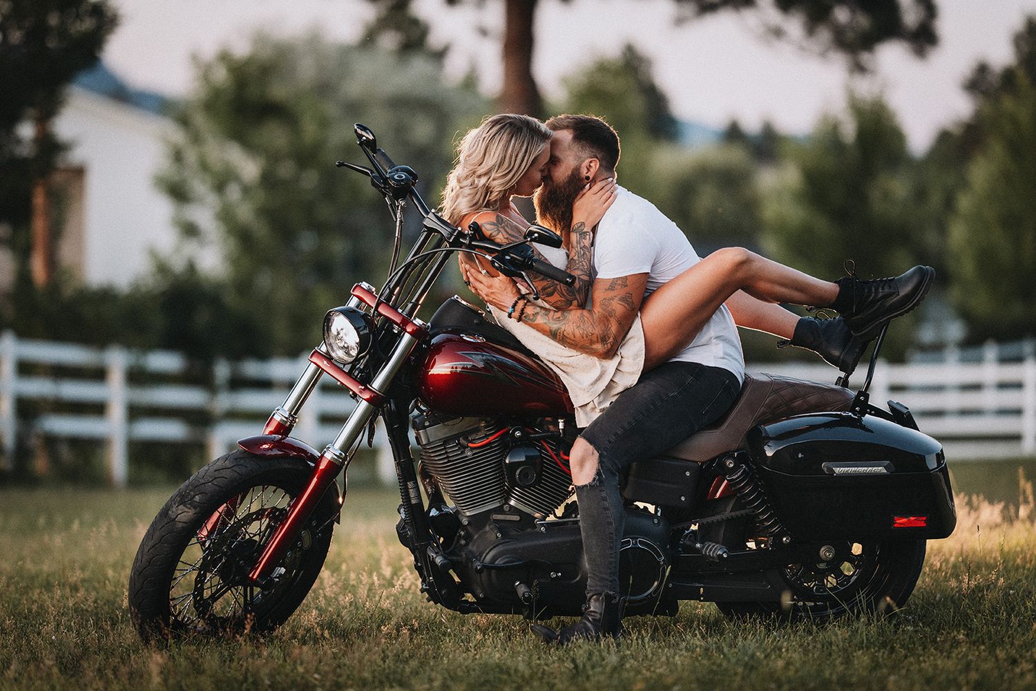 A couple shares an intimate moment while sitting on a parked motorcycle in front of a white fence at sunset.