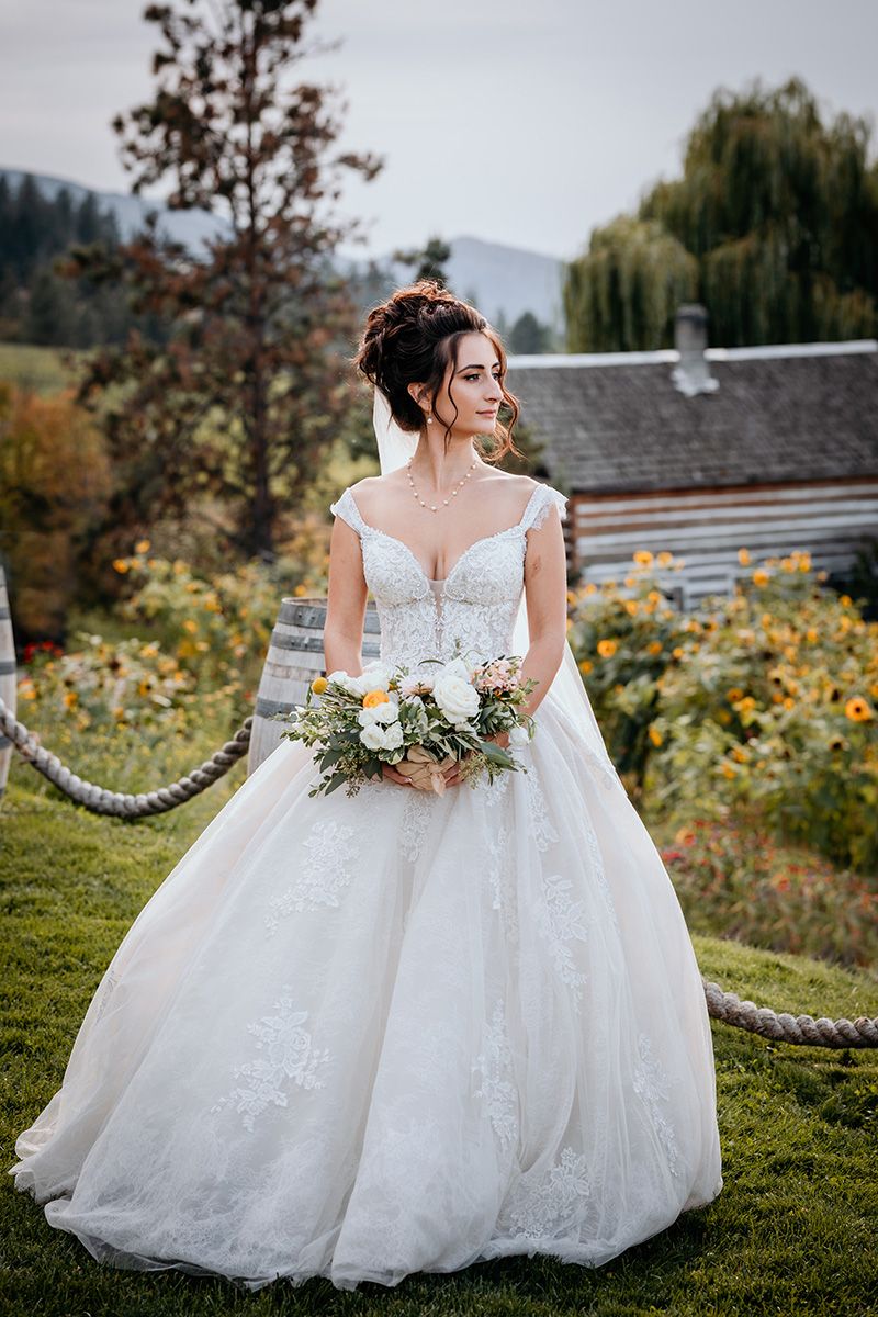 A bride in a flowing white lace gown holds a rustic bouquet while standing on a grassy hill with an old barn in the background.