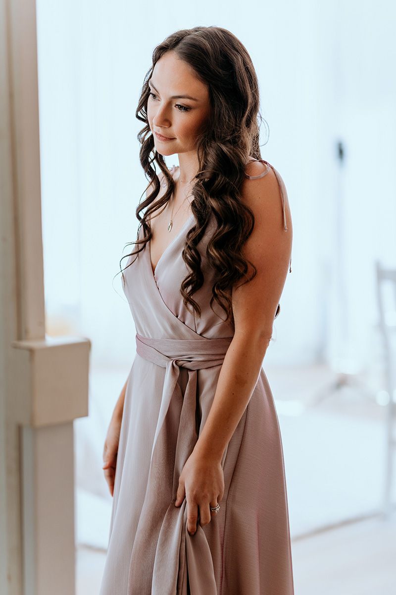 Woman in flowing mauve bridesmaid dress poses elegantly in bright interior setting.