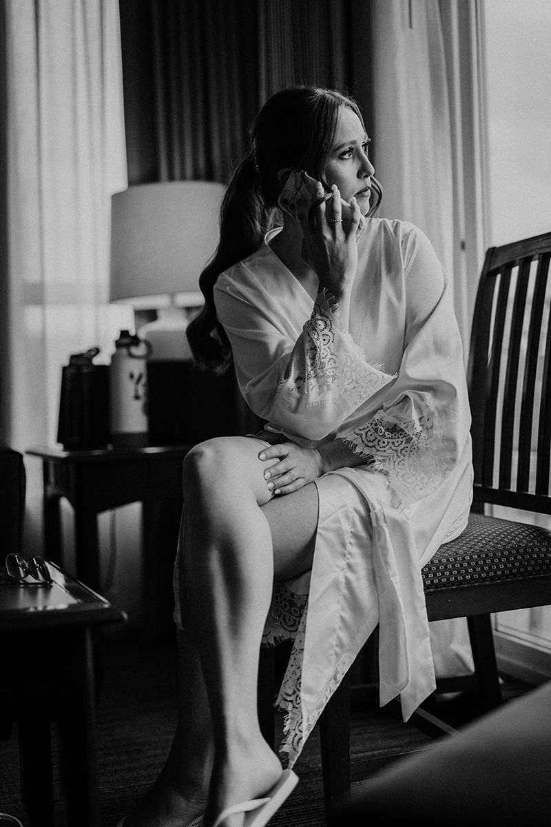 Black and white photo of person in white robe sitting thoughtfully on chair by window in elegant boudoir setting.