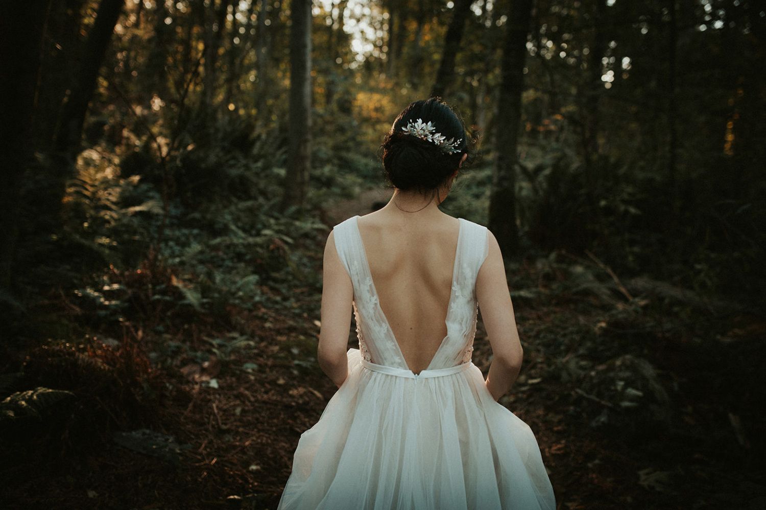 A bride in a white flowy dress walks away from the camera in a moody forest setting during golden hour.