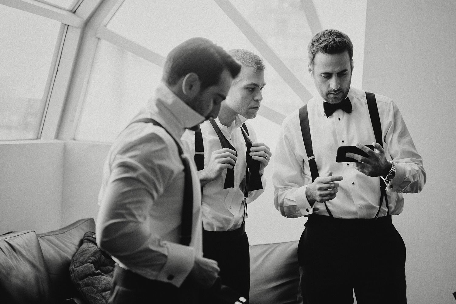 Black and white photo of groomsmen watching a video on a phone on how to tie their bow ties as they get ready for a wedding ceremony.