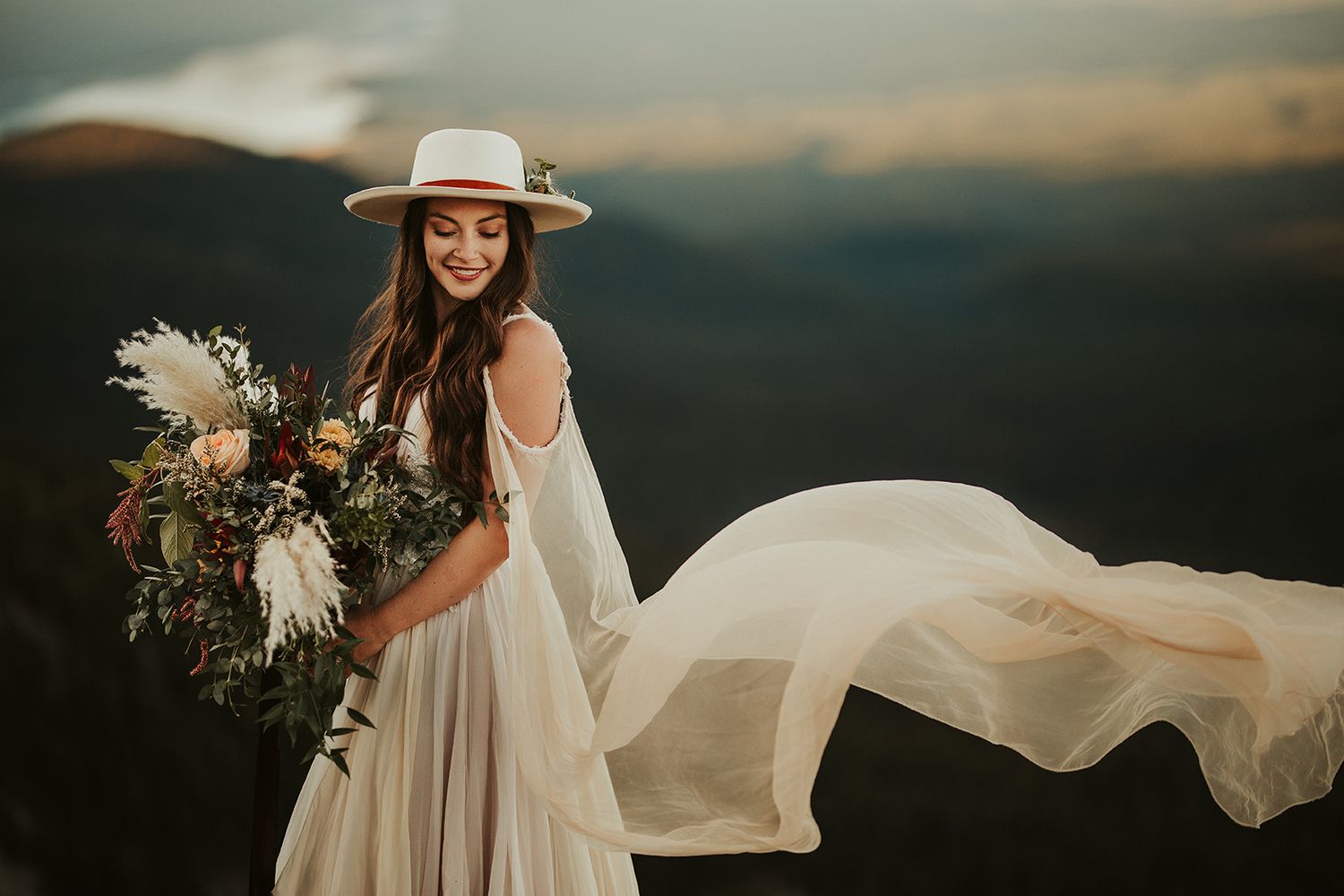 A bride in a flowing white dress poses against a dramatic mountain landscape at sunset with a rustic bouquet.