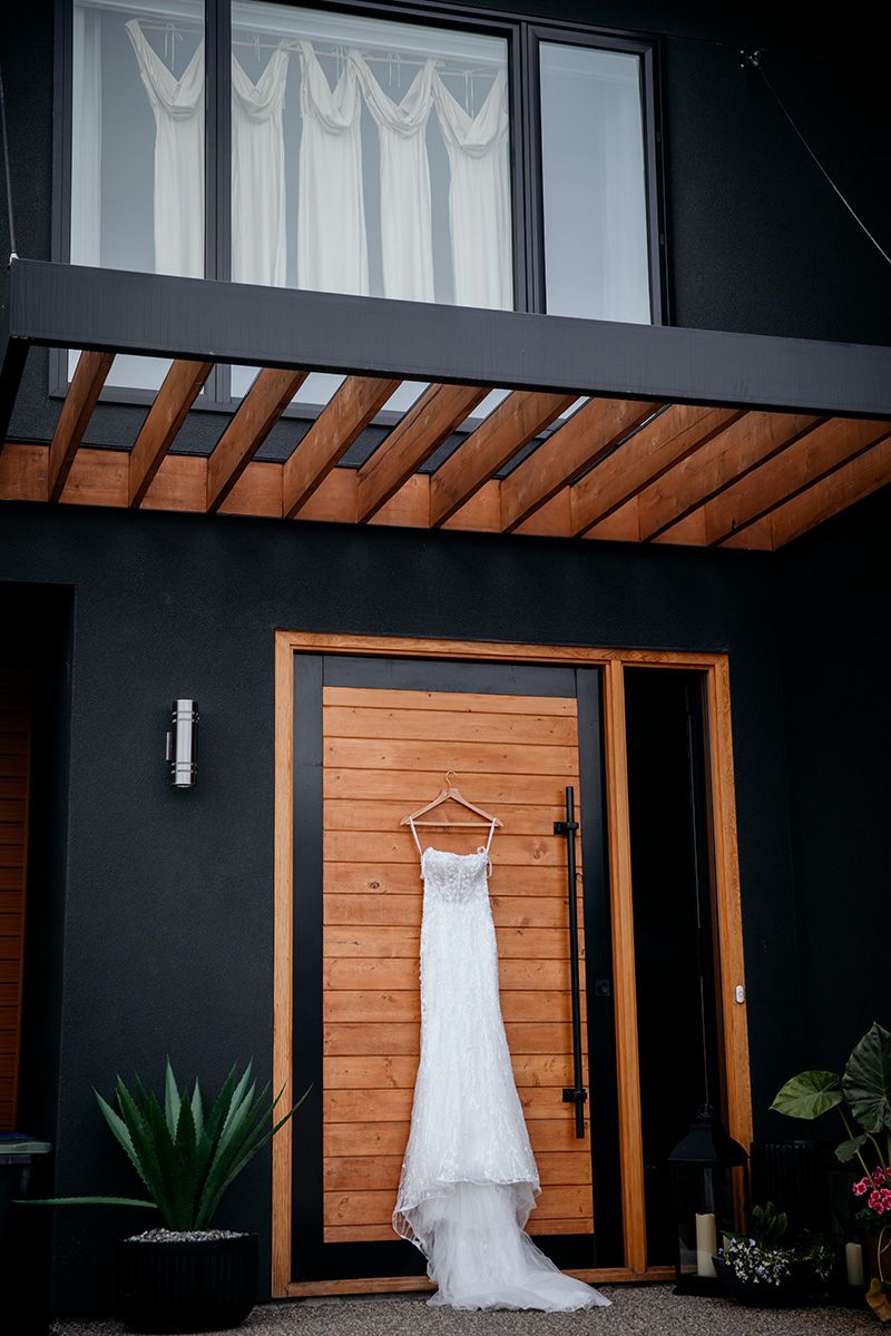 A white wedding dress hangs elegantly against a modern dark wall with wooden accents and tropical plants.