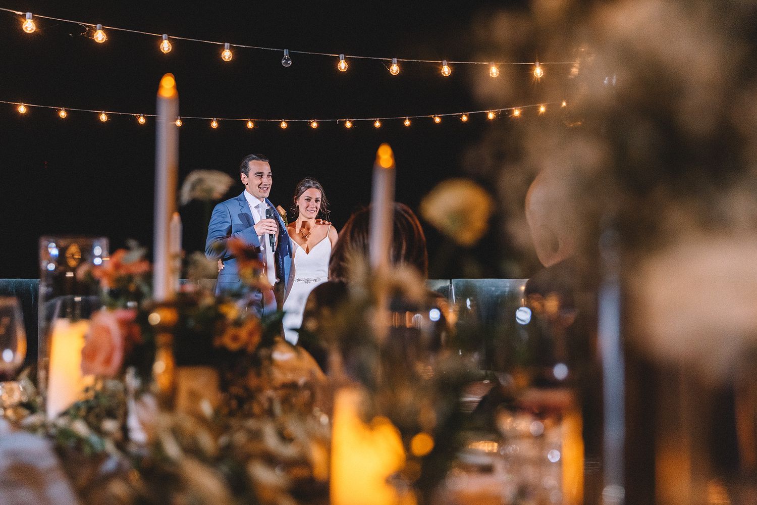 Evening outdoor reception with string lights and candles creating a warm romantic atmosphere during a wedding couple's speech..