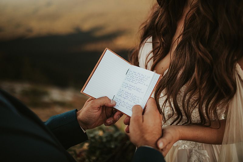 A groom in a blue suit reading handwritten vows during an outdoor during golden hour wedding ceremony.