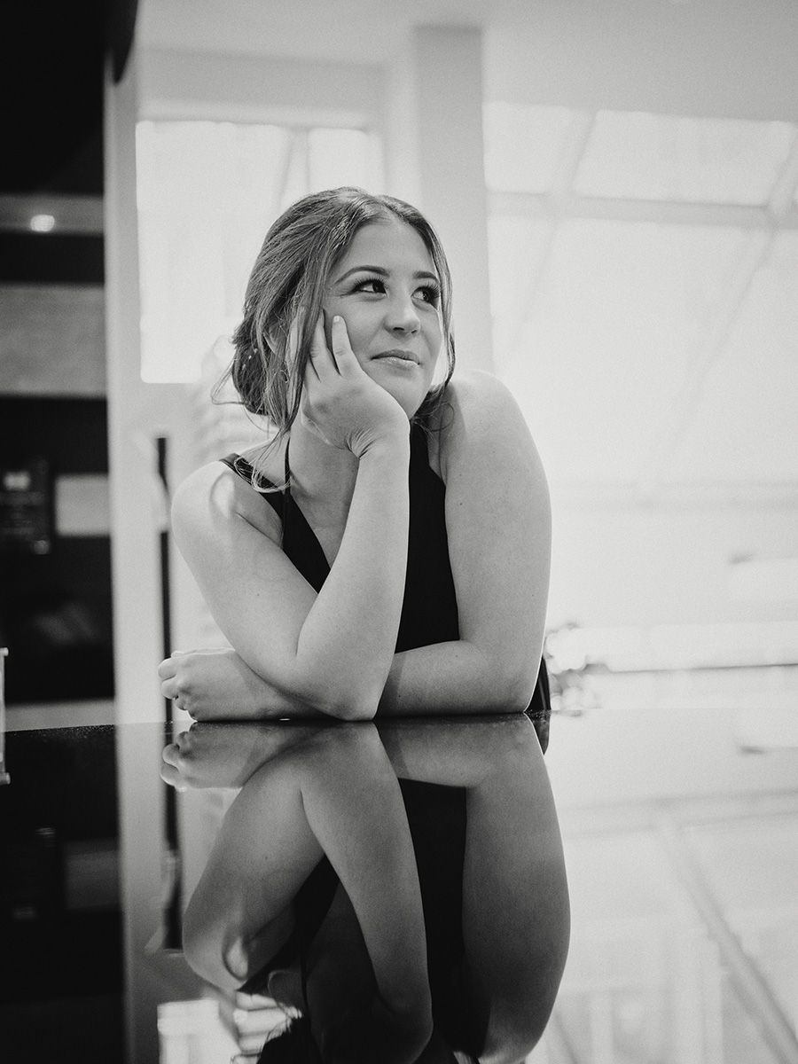 Black and white portrait of a woman sitting in a contemplative pose with hand resting on chin in an indoor setting.