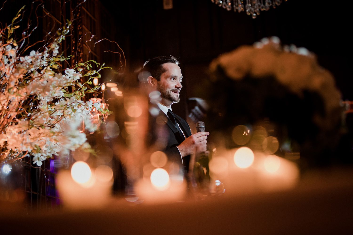 A man gives a speech at a romantic wedding reception with glowing string lights creating a warm, intimate atmosphere.