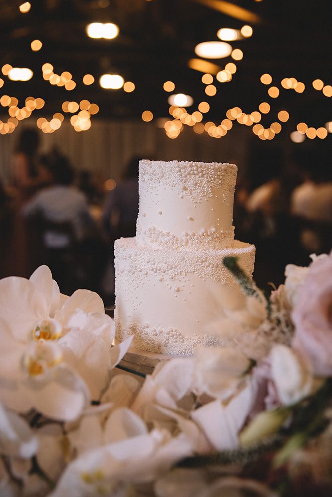 Elegant white wedding cake decorated with delicate pearl details surrounded by soft white flowers and twinkling bokeh lights.