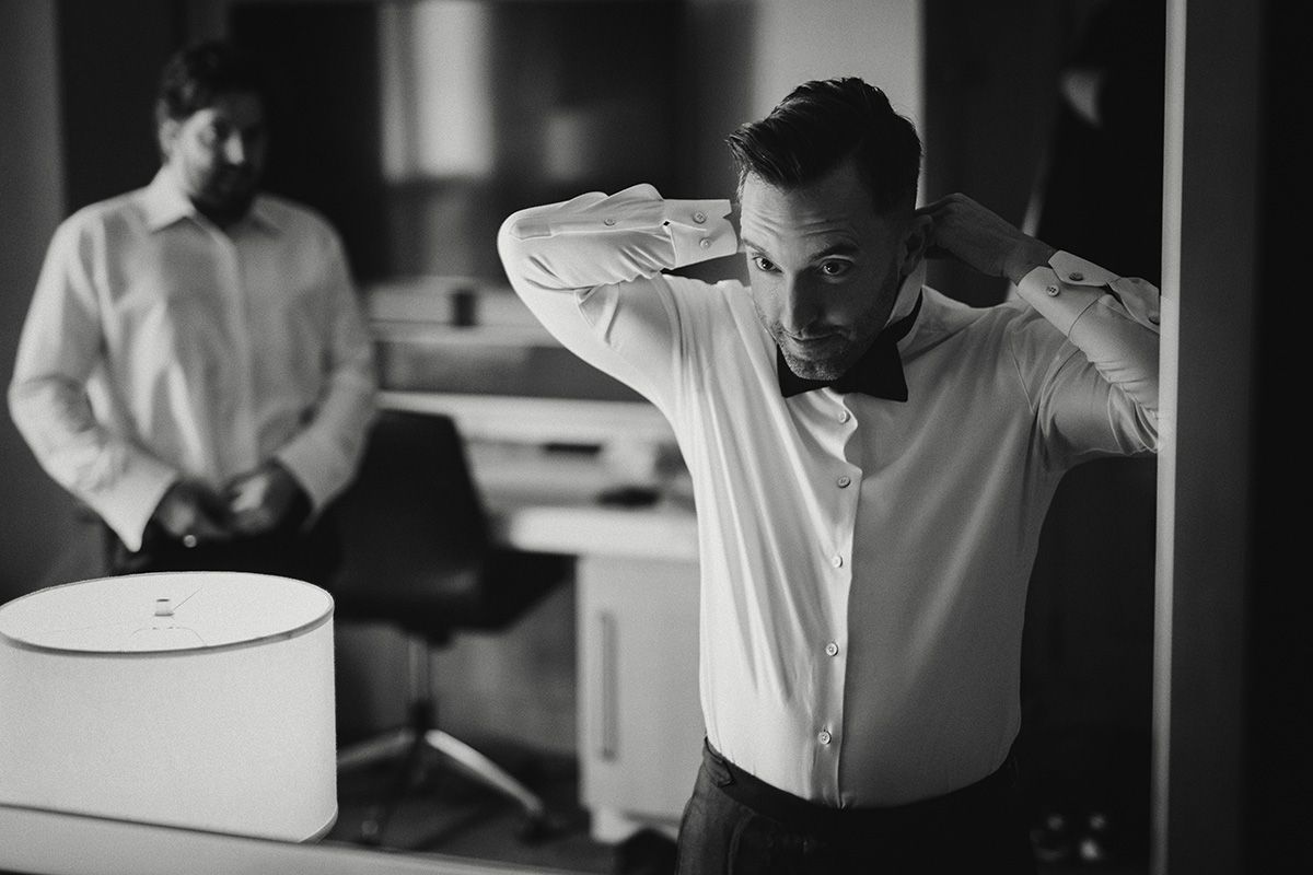 A black and white photo of man in a white dress shirt looking in a mirror while he ties his bow-tie.