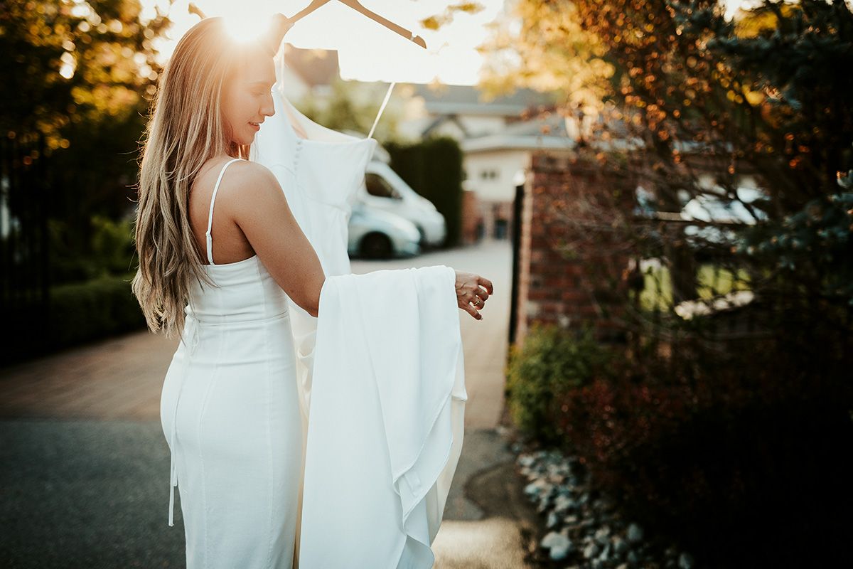 Sunlit golden hour photoshoot of a bride in a flowing white night dress walking through a garden setting holding her wedding dress.