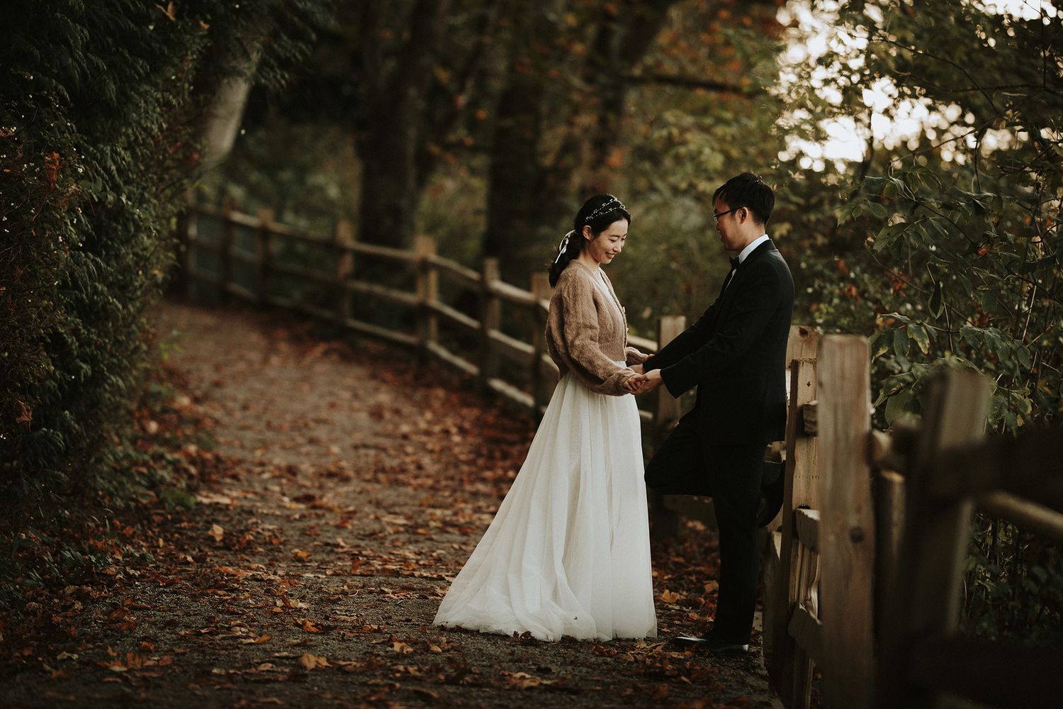 A romantic autumn wedding scene with a couple standing on a leaf-covered path beside a rustic wooden fence.