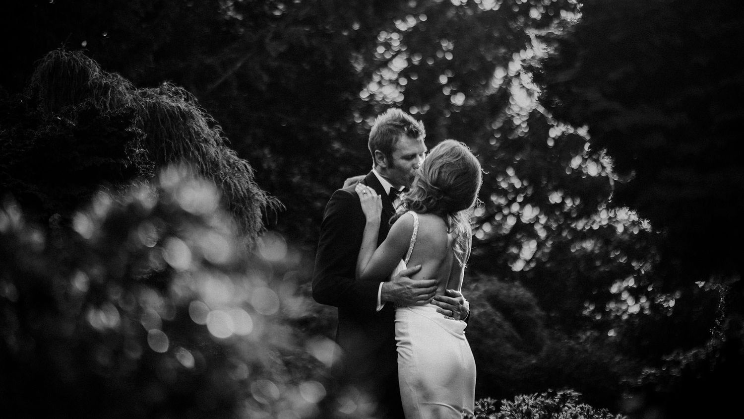 A black and white wedding photograph showing romantic moments between a groom in a bow-tie kissing his bride in an outdoor setting.