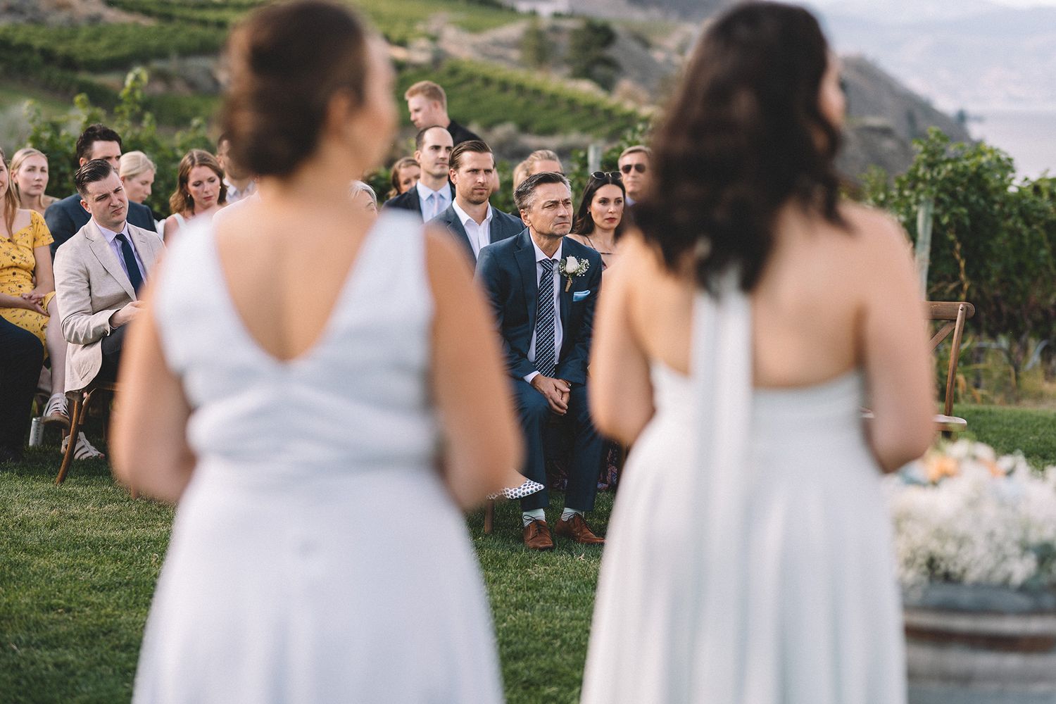 Wedding guests smile as they watch two bridesmaids in white baby blue dresses during an outdoor ceremony in a scenic hillside vineyard.