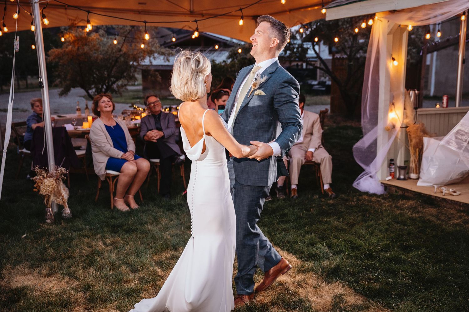 A bride and groom dancing together at their outdoor evening wedding reception under a white tent with warm lighting.