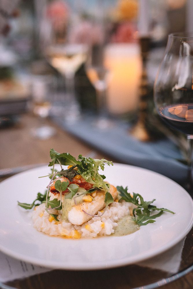 Elegant plated seafood dish with microgreens and risotto served on white china with wine glass in background.
